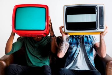 dos personas sentadas en un sillón, en las manos tienen un televisor. Sostienen el aparato a la altura de la cara, tapándose la cabeza. Los televisores tienen colores vibrantes, como rojo, turquesa y amarillo