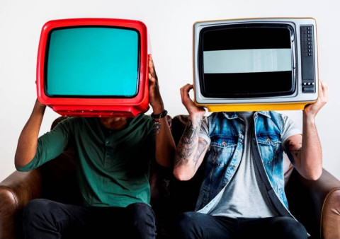 dos personas sentadas en un sillón, en las manos tienen un televisor. Sostienen el aparato a la altura de la cara, tapándose la cabeza. Los televisores tienen colores vibrantes, como rojo, turquesa y amarillo